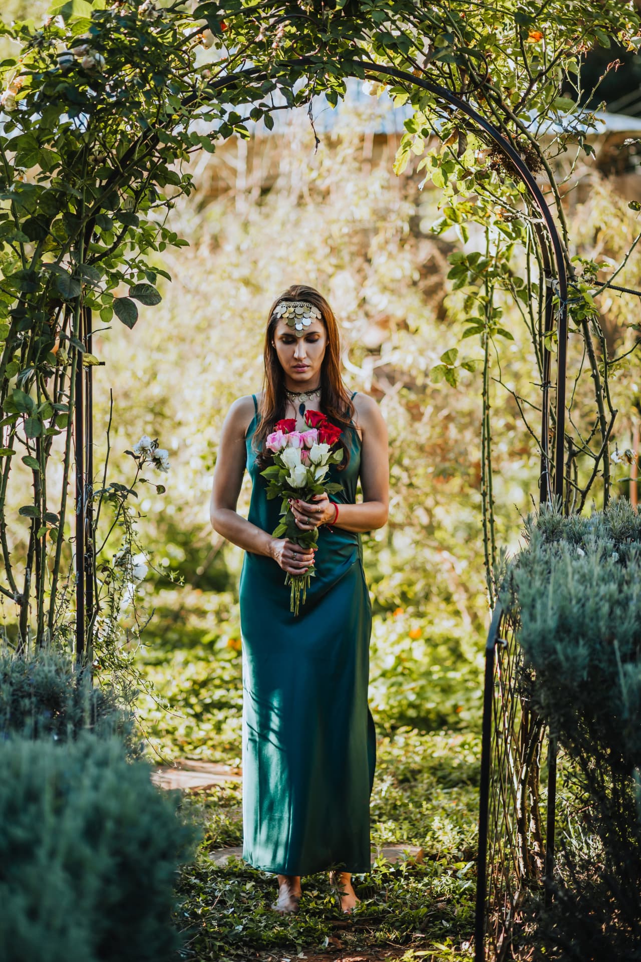 Noa standing in a garden arch wearing emerald silk and holding roses